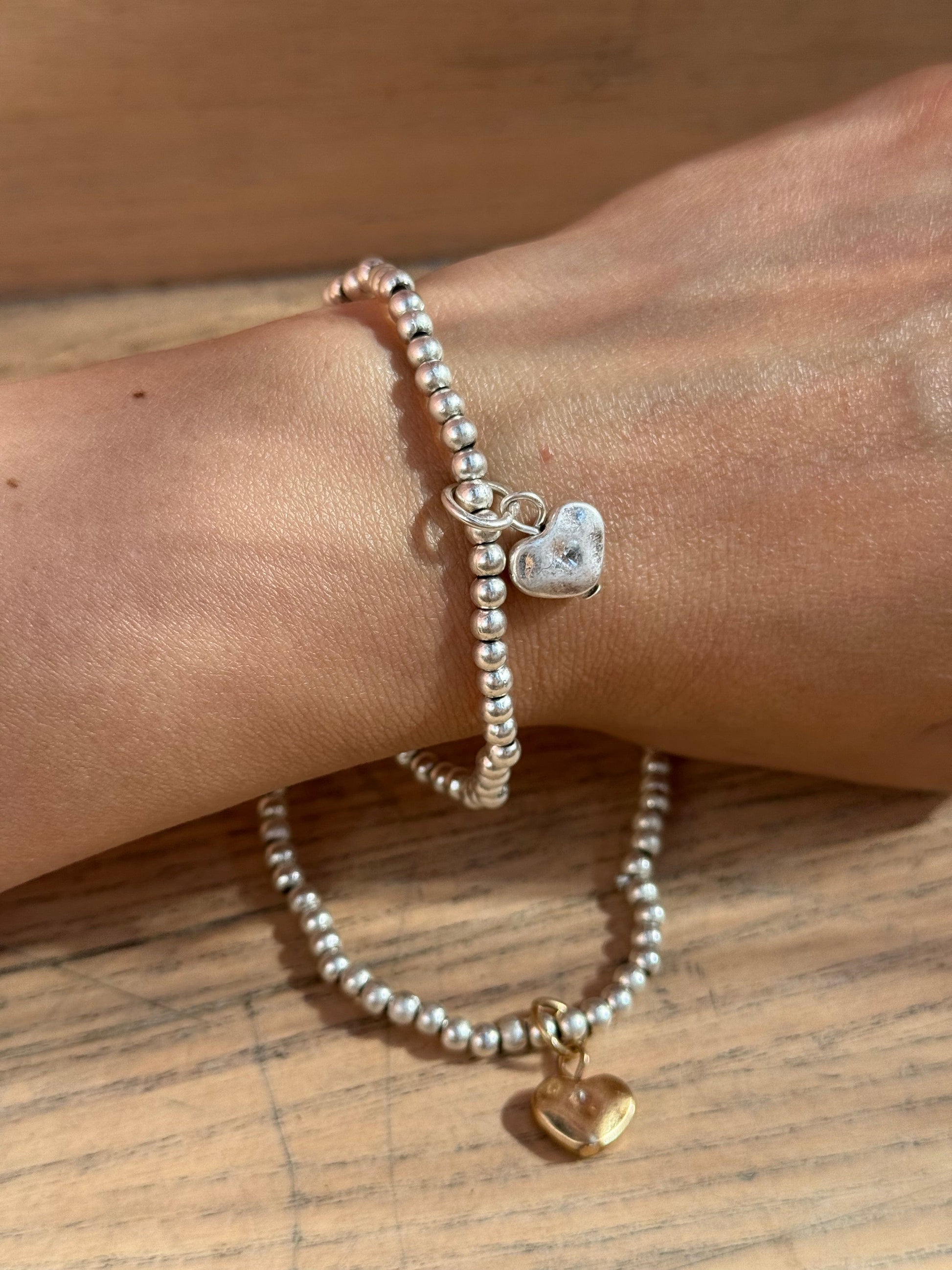 Two beaded silver bracelets with heart charms on a wrist, gold and silver